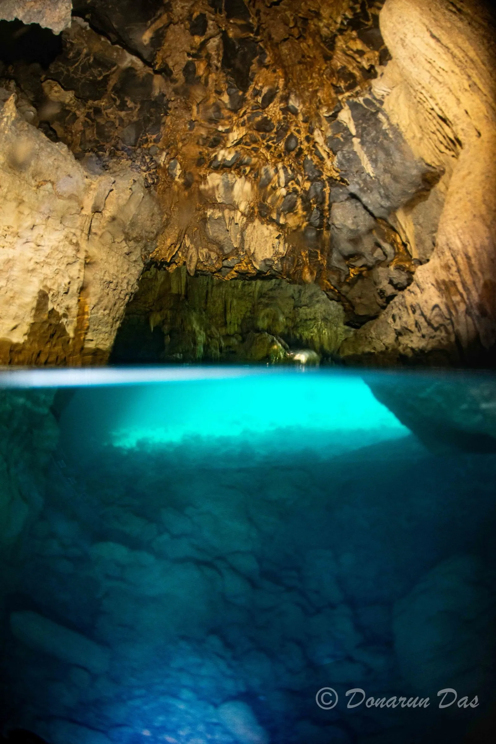 Over-under view inside a Meghalaya cavern: stalactite-hung limestone ceiling above a daylight-blue flooded passage