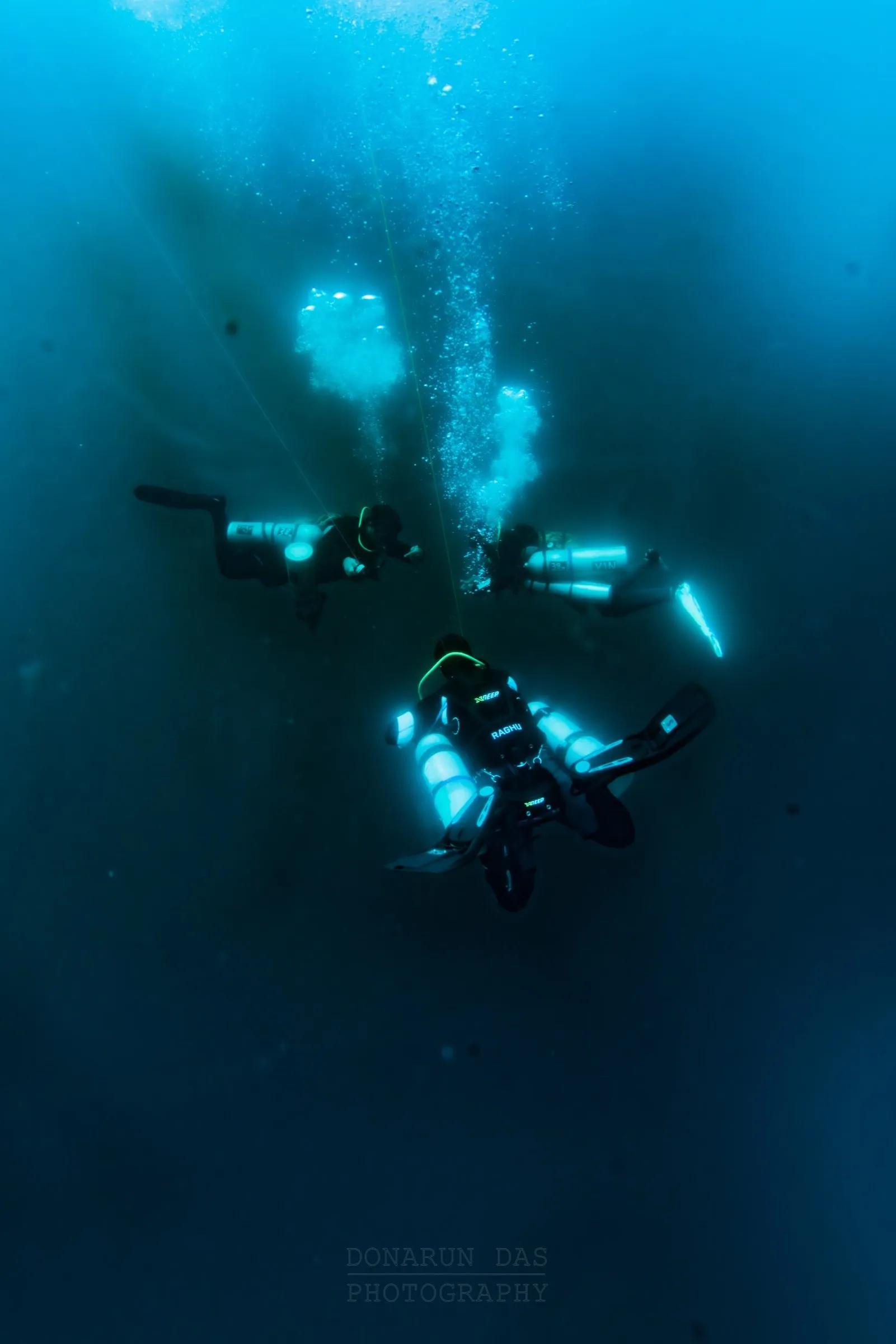 Three technical divers descending a shotline into deep blue water, two carrying stage cylinders