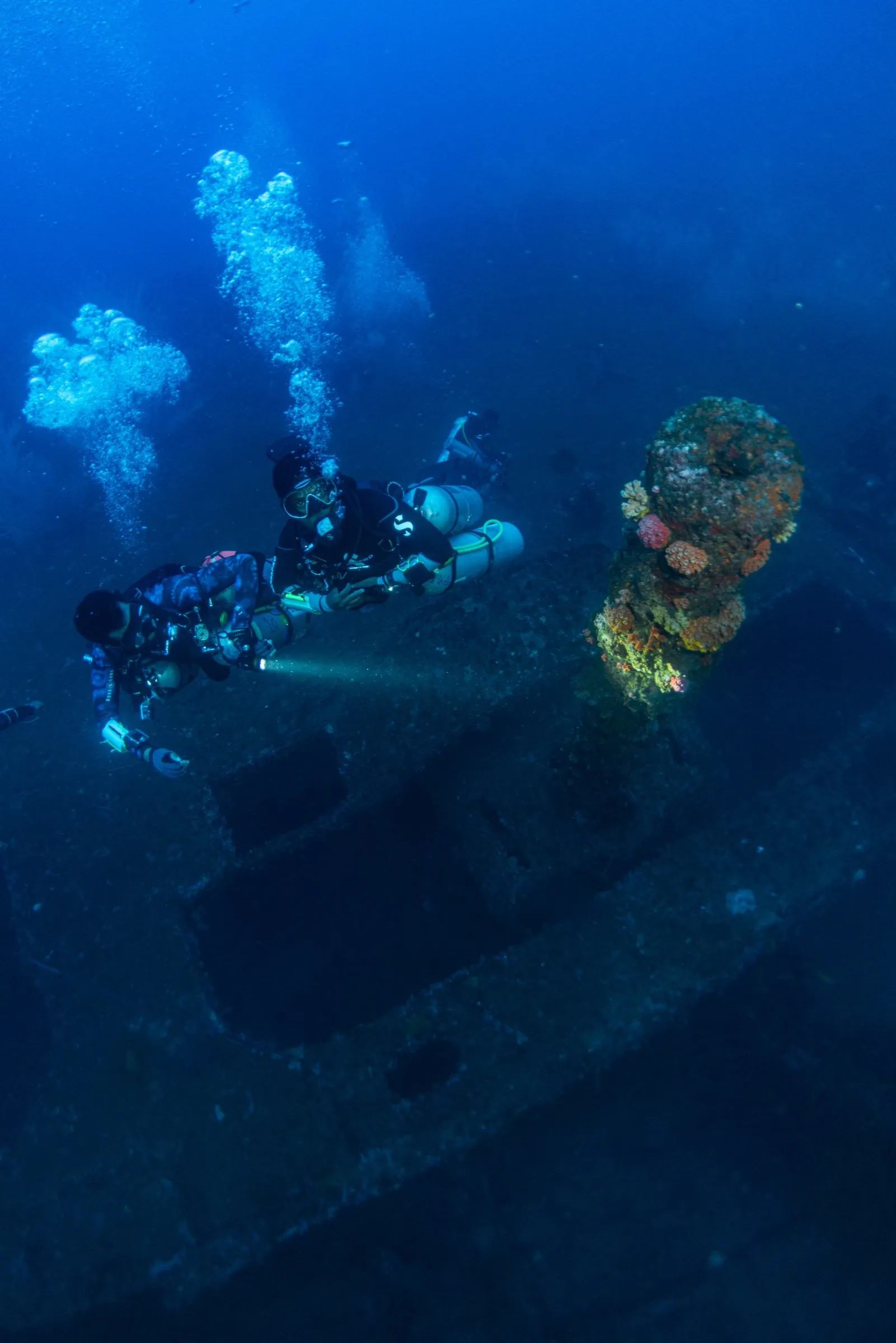 Two technical divers over a coral-grown wreck, one in sidemount and one in backmount, lights on and bubbles rising