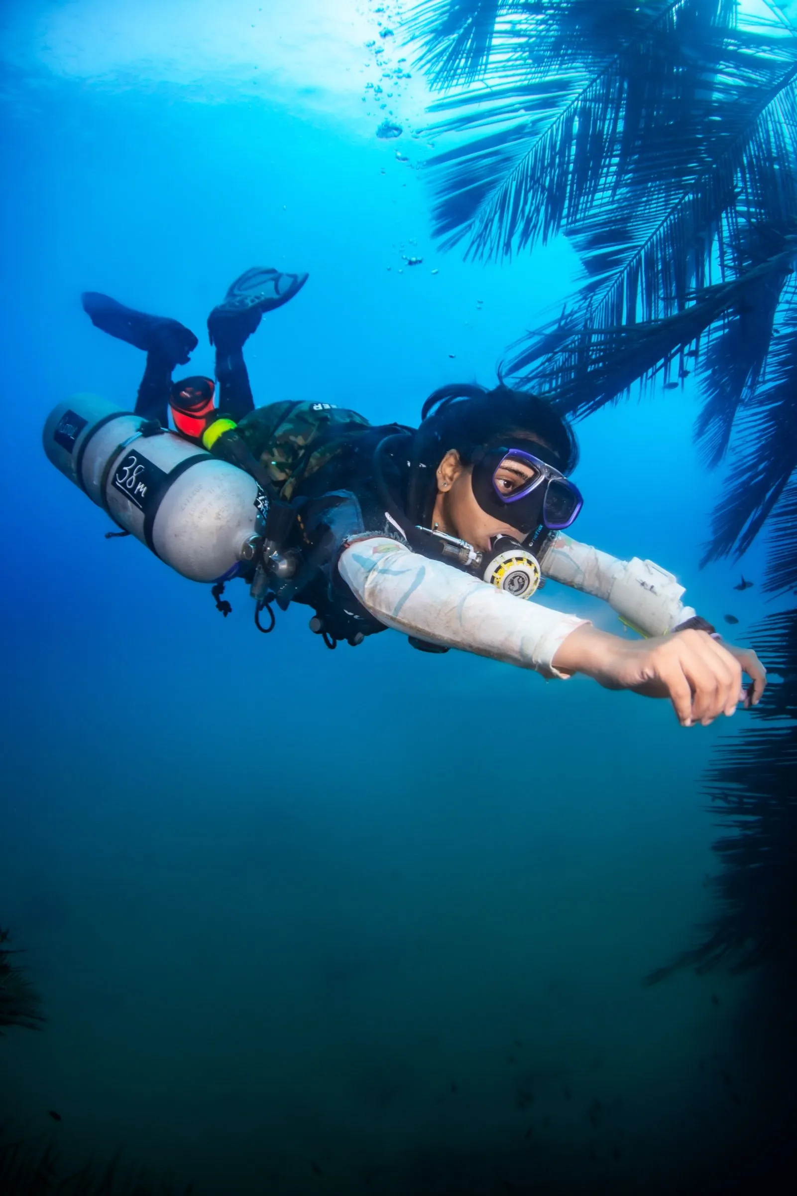 Sidemount diver in deep blue water, single cylinder marked 38m clipped along the body, palm fronds silhouetted at the surface above