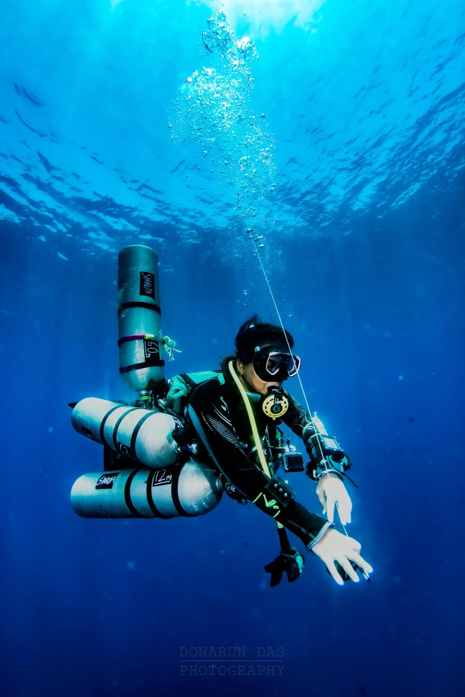 Sidemount technical diver hanging on an upline in clear blue water with stage cylinders during ascent