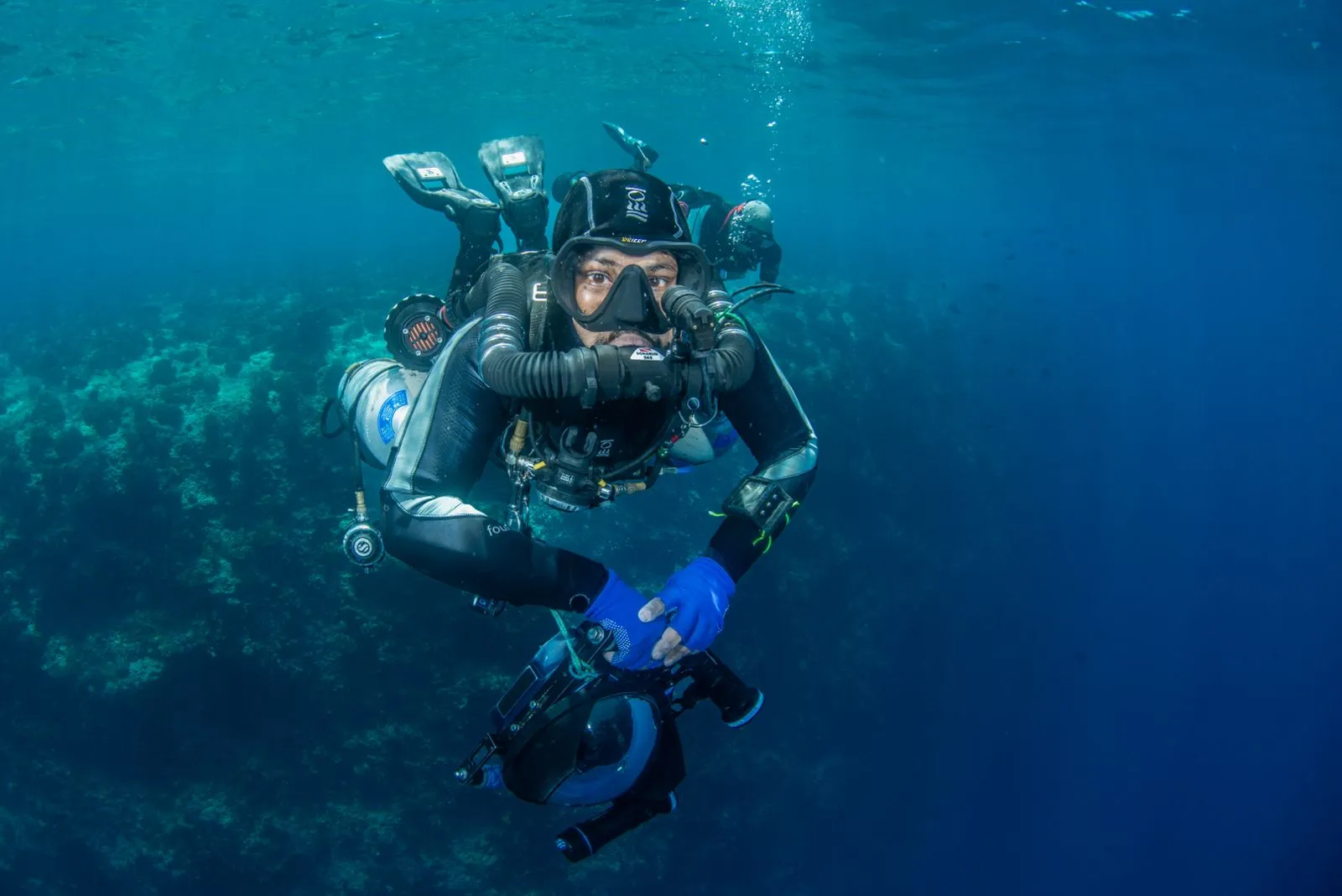 Diver on a chest-mounted KISS Sidewinder rebreather holding a DPV in blue water above a reef, corrugated loop hoses across the chest