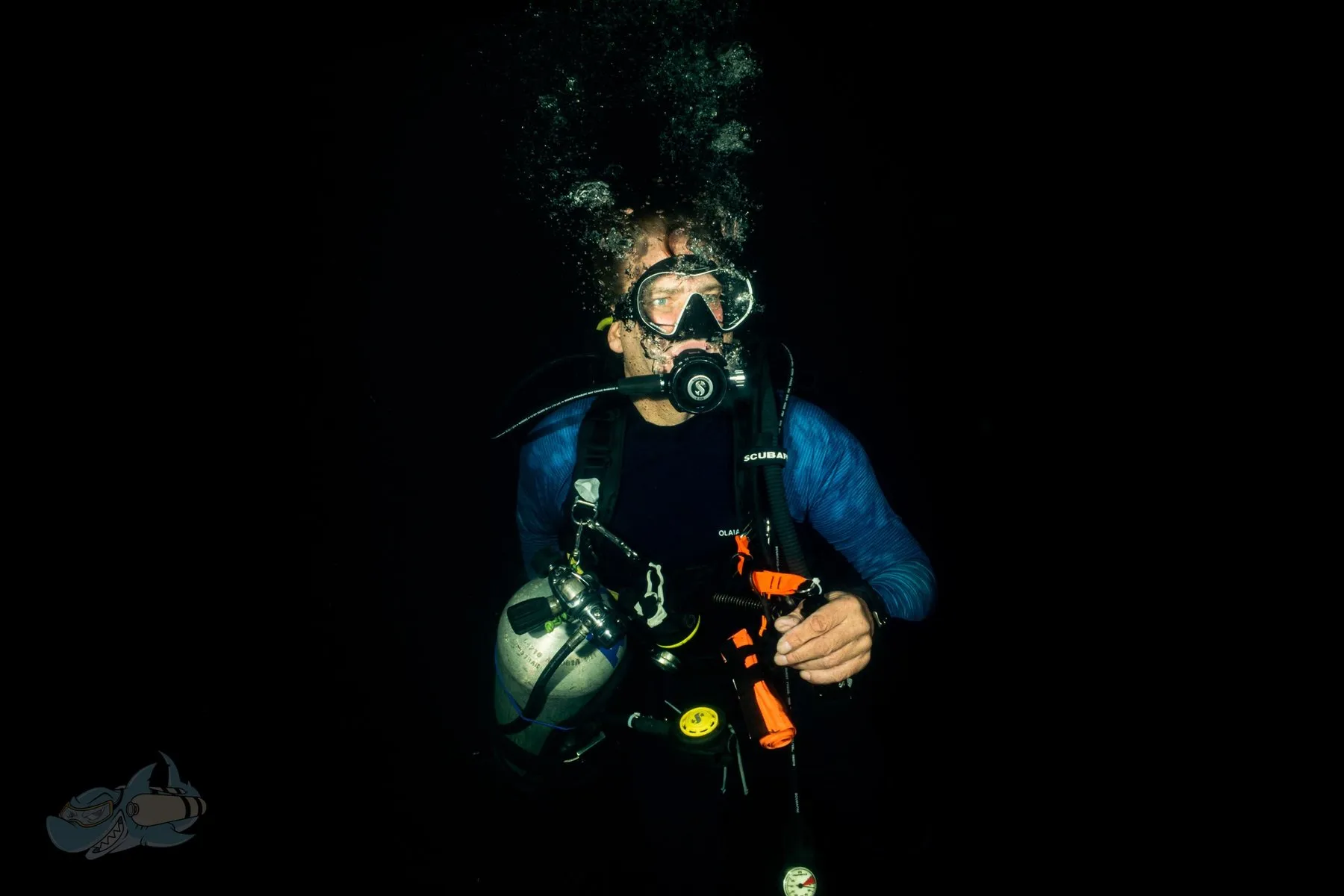 Single backmount diver lit against black water at depth, holding a deployed surface marker and gauges