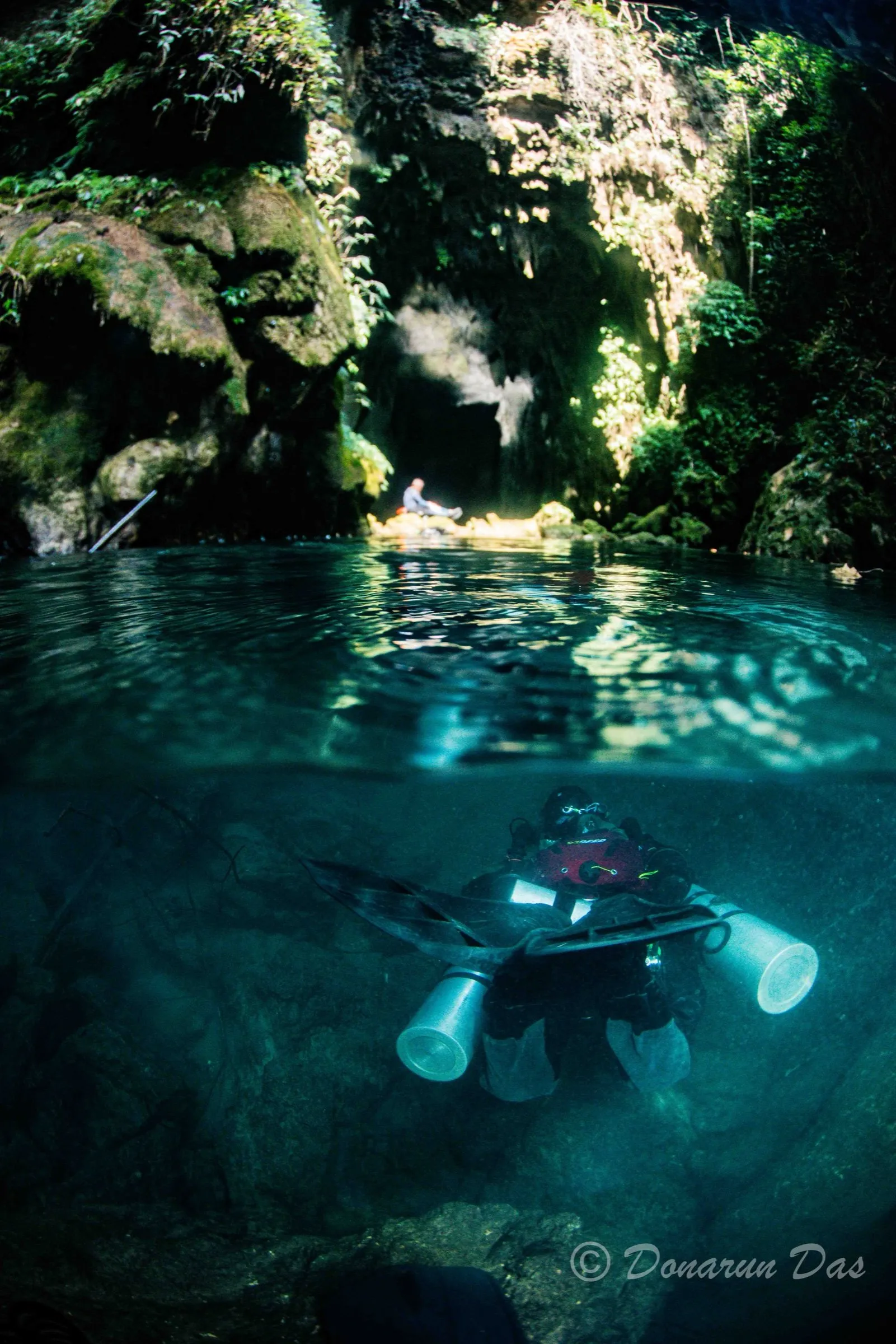 Split over-under view of a rocky daylit cavern entrance with a diver on a DPV in the clear water below