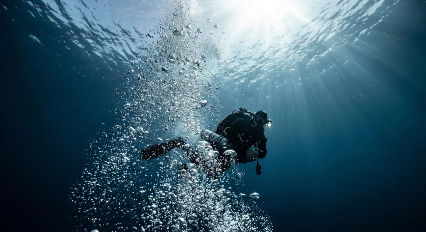 A technical scuba diver ascending through a rising column of silver bubbles toward the sunlit surface in deep blue water