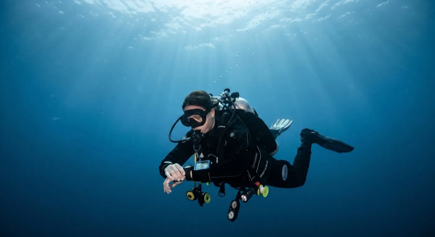 A technical diver hovering mid-water on a slow ascent, checking a wrist dive computer, with shafts of sunlight coming from the surface above