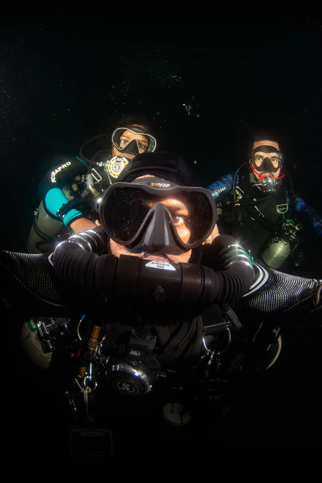 Three divers in dark water: Donarun Das on a chest-mounted CCR in the foreground, two open-circuit backmount divers behind