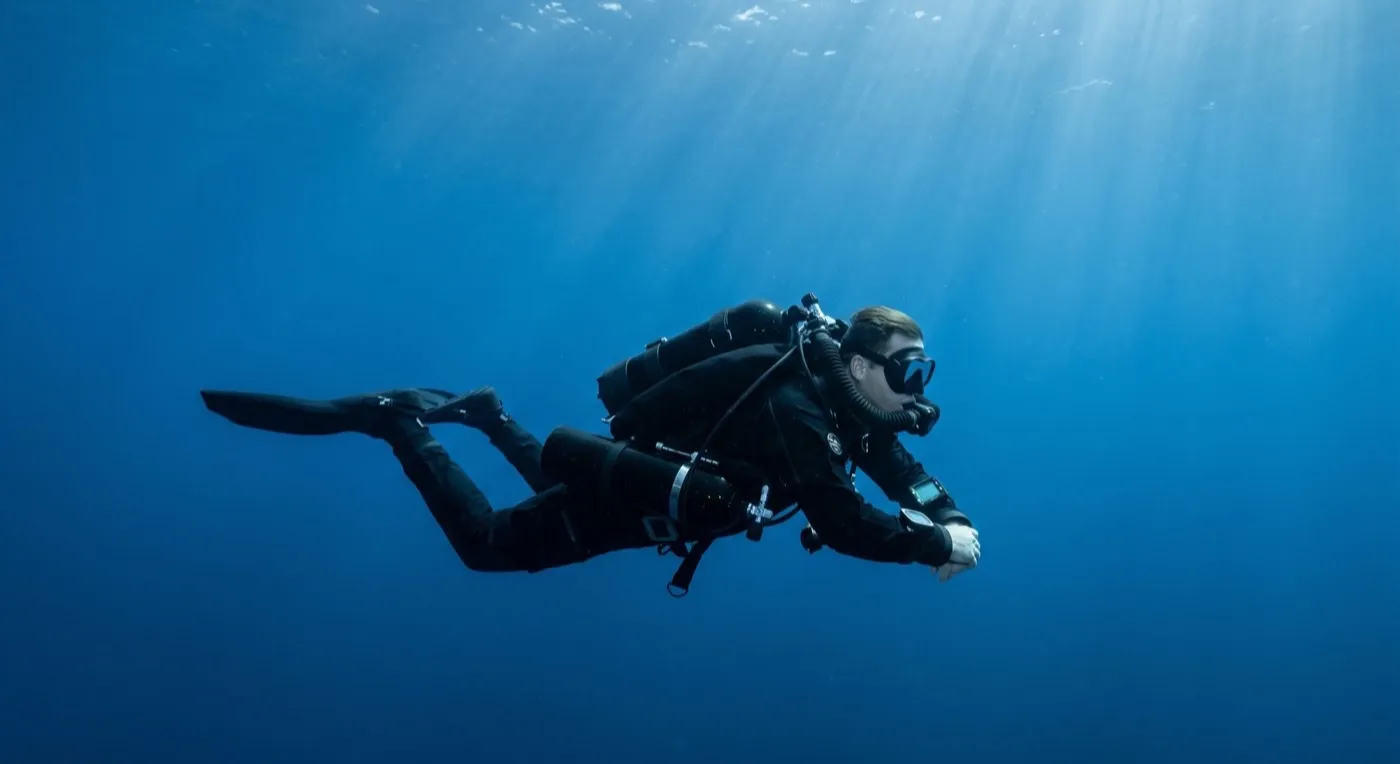 A technical diver gliding mid-water on a sleek black sidemount closed-circuit rebreather with no exhaust bubbles, in deep blue water