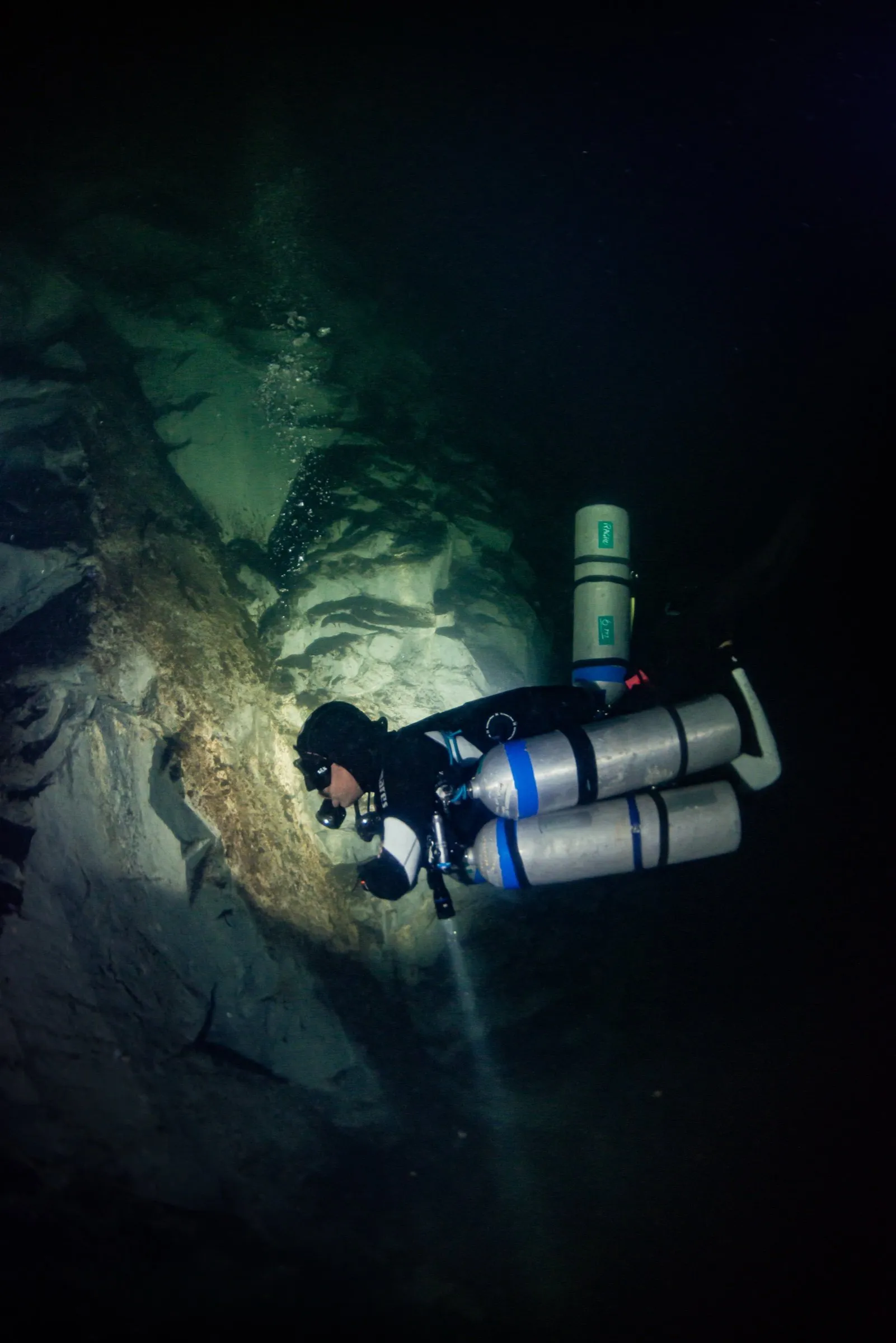 Sidemount technical diver with multiple side-slung cylinders and a primary light inside a dark rock cavern