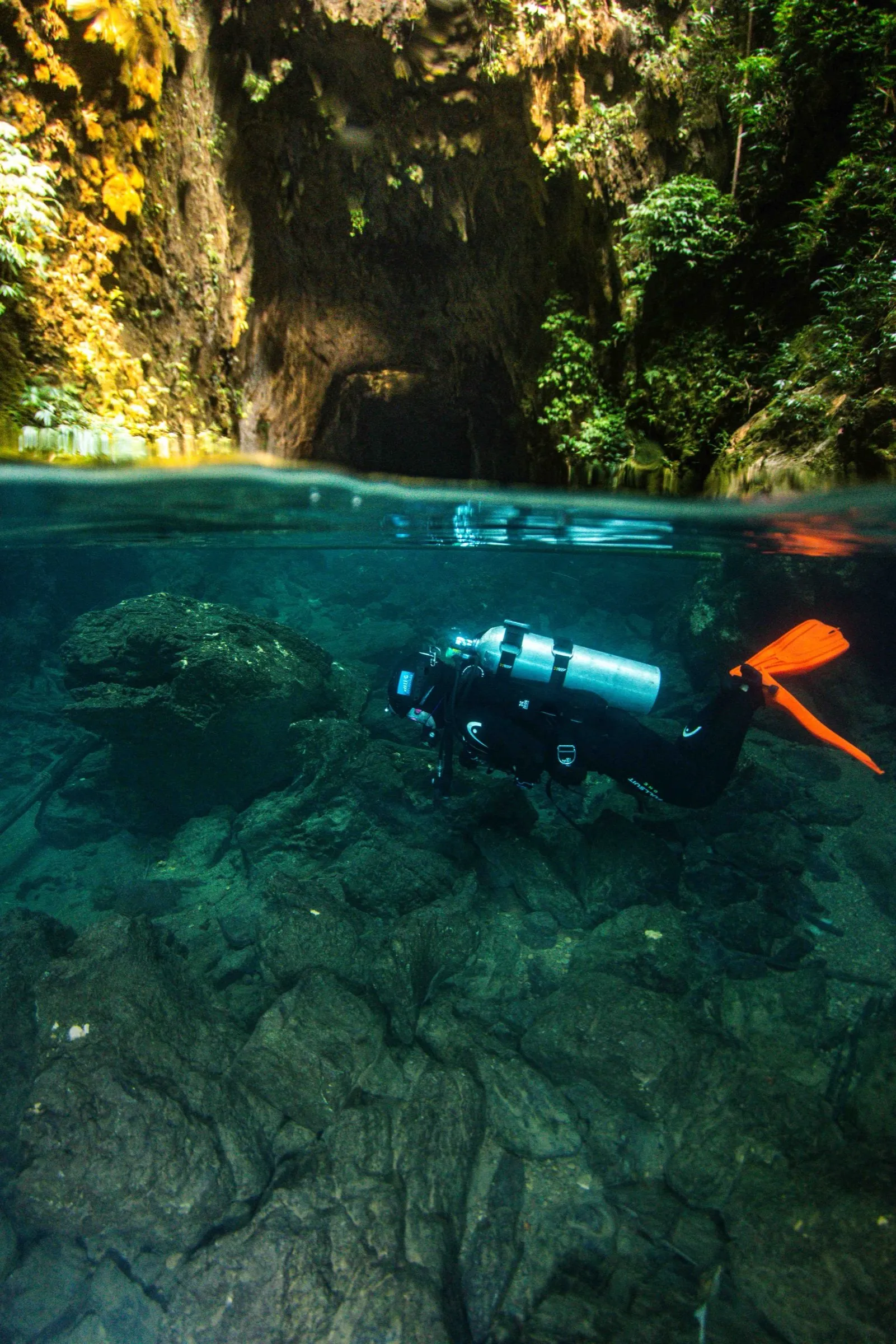 Sidemount diver in horizontal trim just below the surface at a rocky cavern mouth, daylight visible above