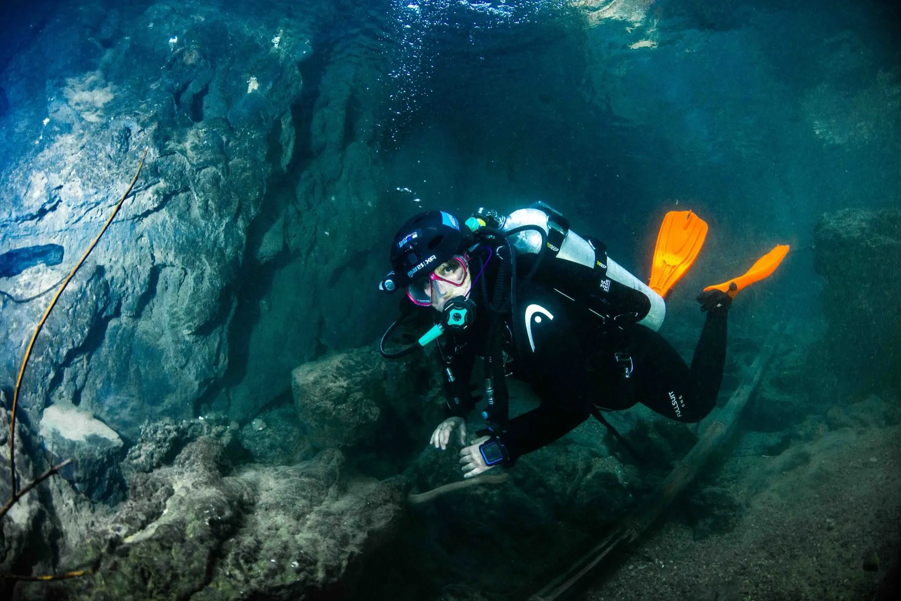 Diver in black wetsuit and orange fins holding flat horizontal trim with one hand steadied on rock in a silty cavern