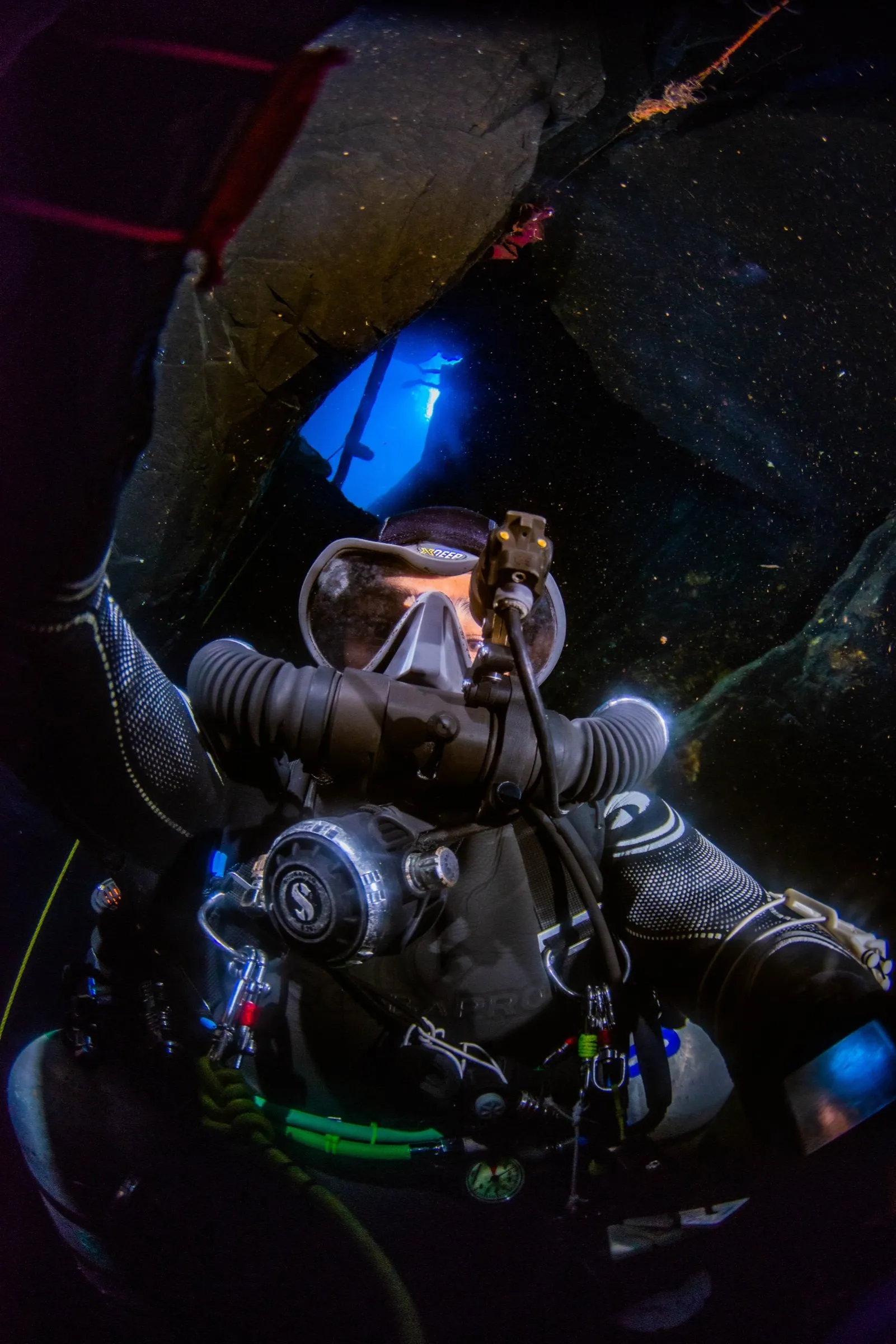 Diver on a chest-mounted KISS Sidewinder rebreather in a dark overhead environment, counterlungs and loop hoses lit against the rock
