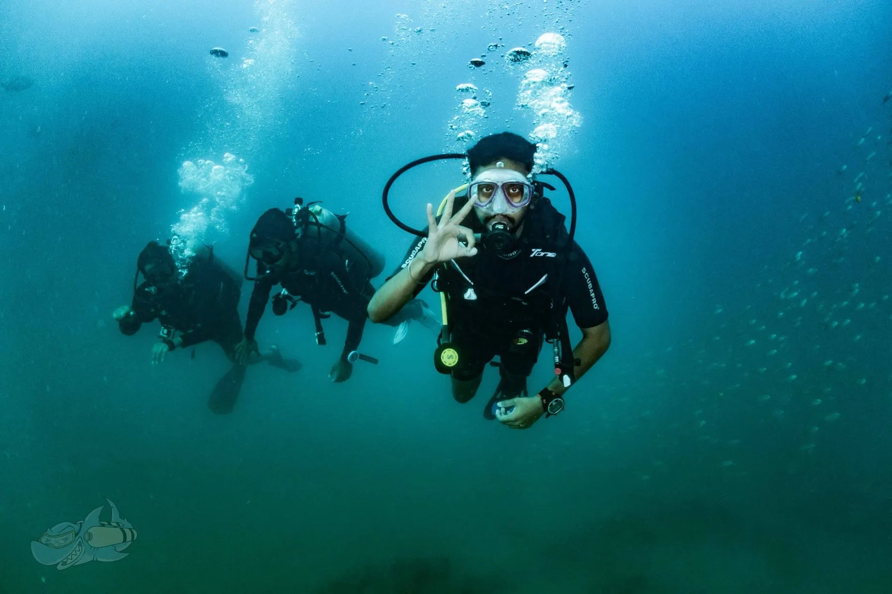 Three-diver buddy team in midwater, the lead diver facing the camera with an OK hand signal as bubbles rise