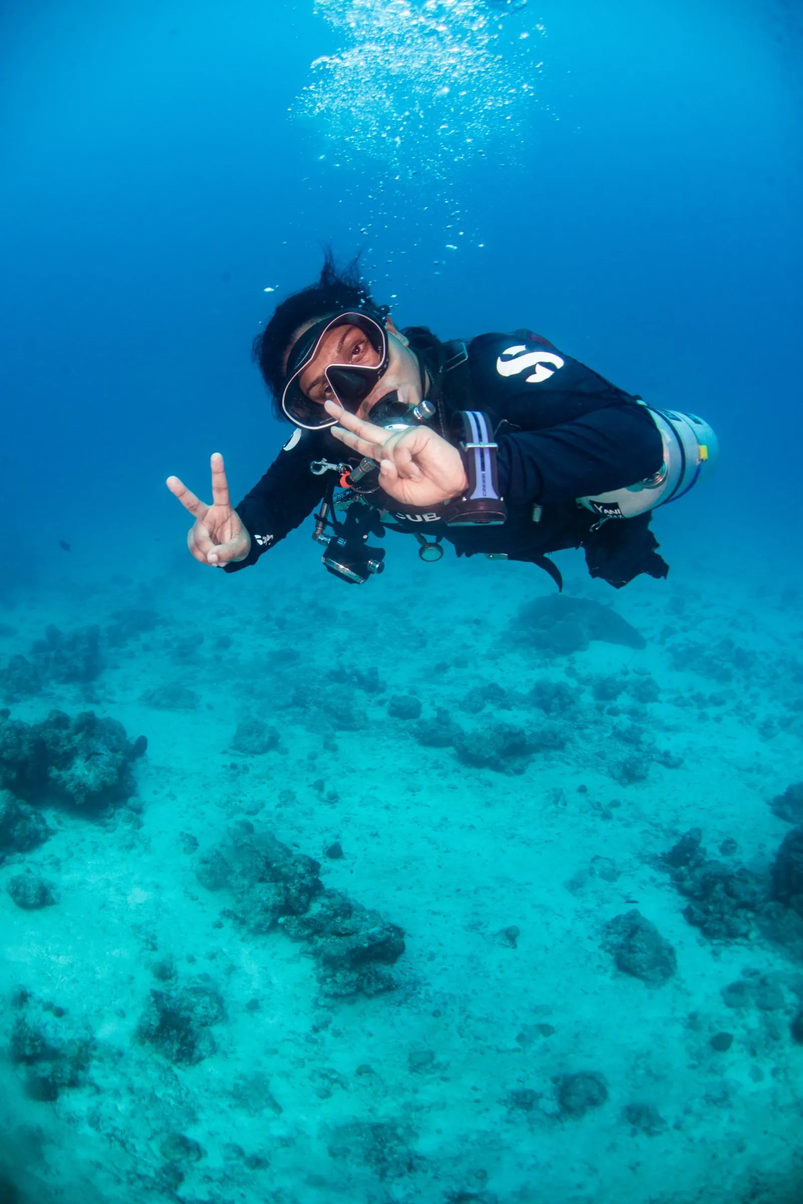 Sidemount diver hovering over a reef holding a compact camera, flashing peace signs to the lens, single side cylinder