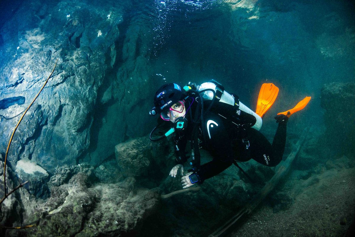 Diver in orange fins holding flat horizontal trim with a hand steadied on rock in a silty cavern