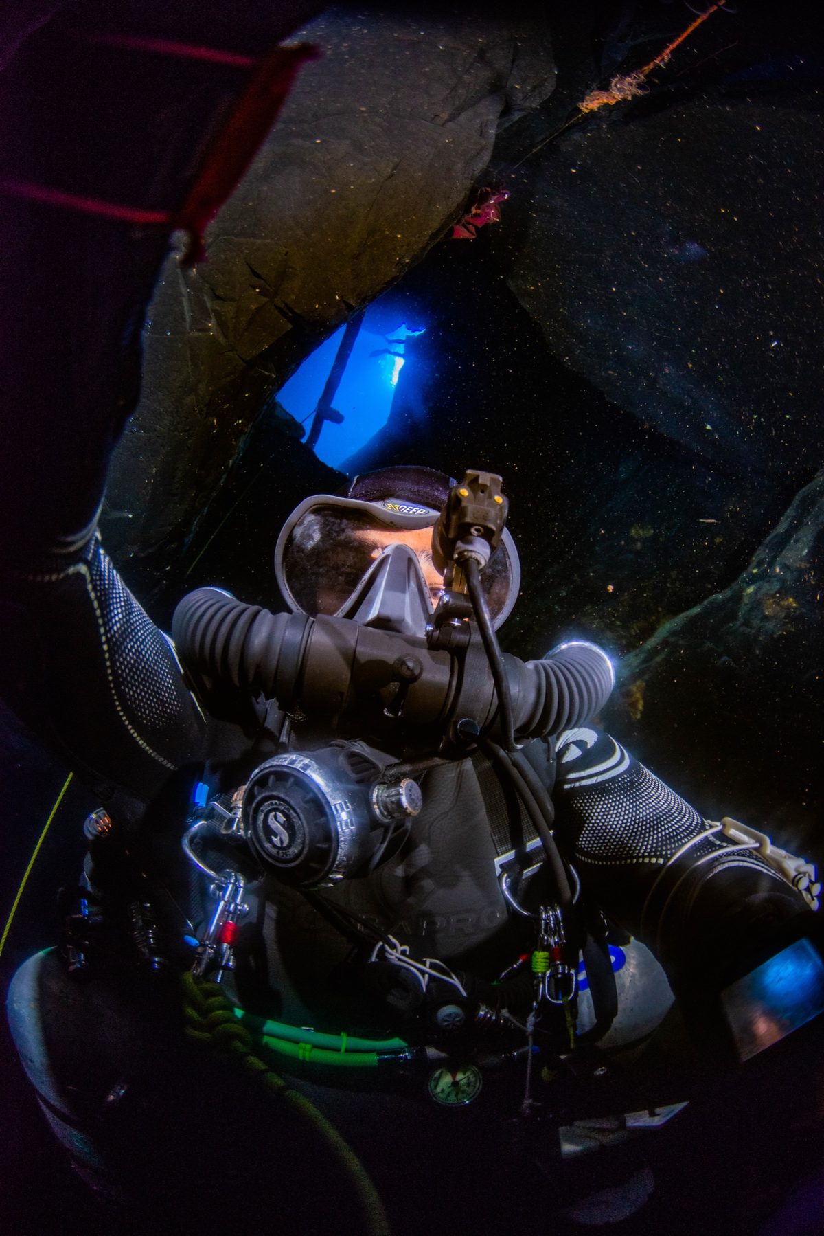 Diver on a chest-mounted KISS Sidewinder rebreather in a dark overhead environment, counterlungs and loop hoses lit against the rock