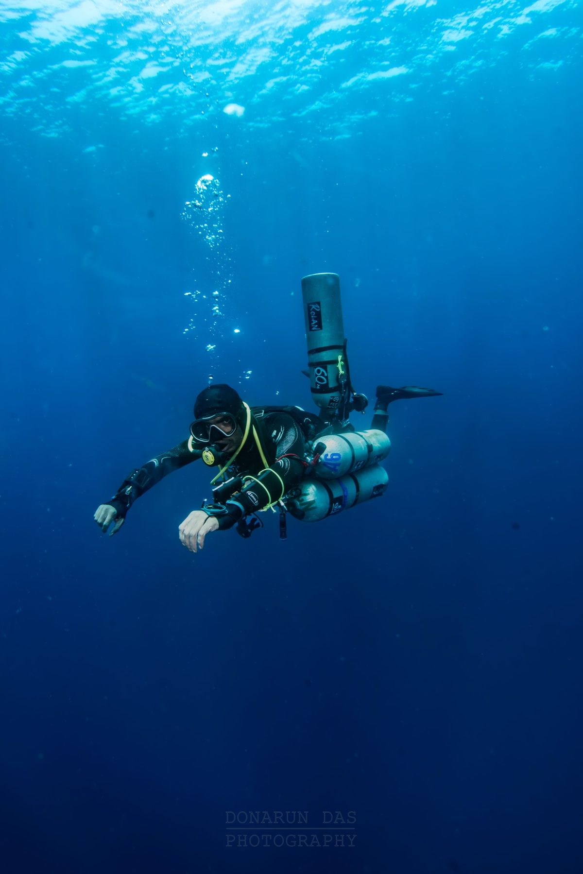 Sidemount technical diver suspended in open blue water holding a decompression stop with stage cylinders