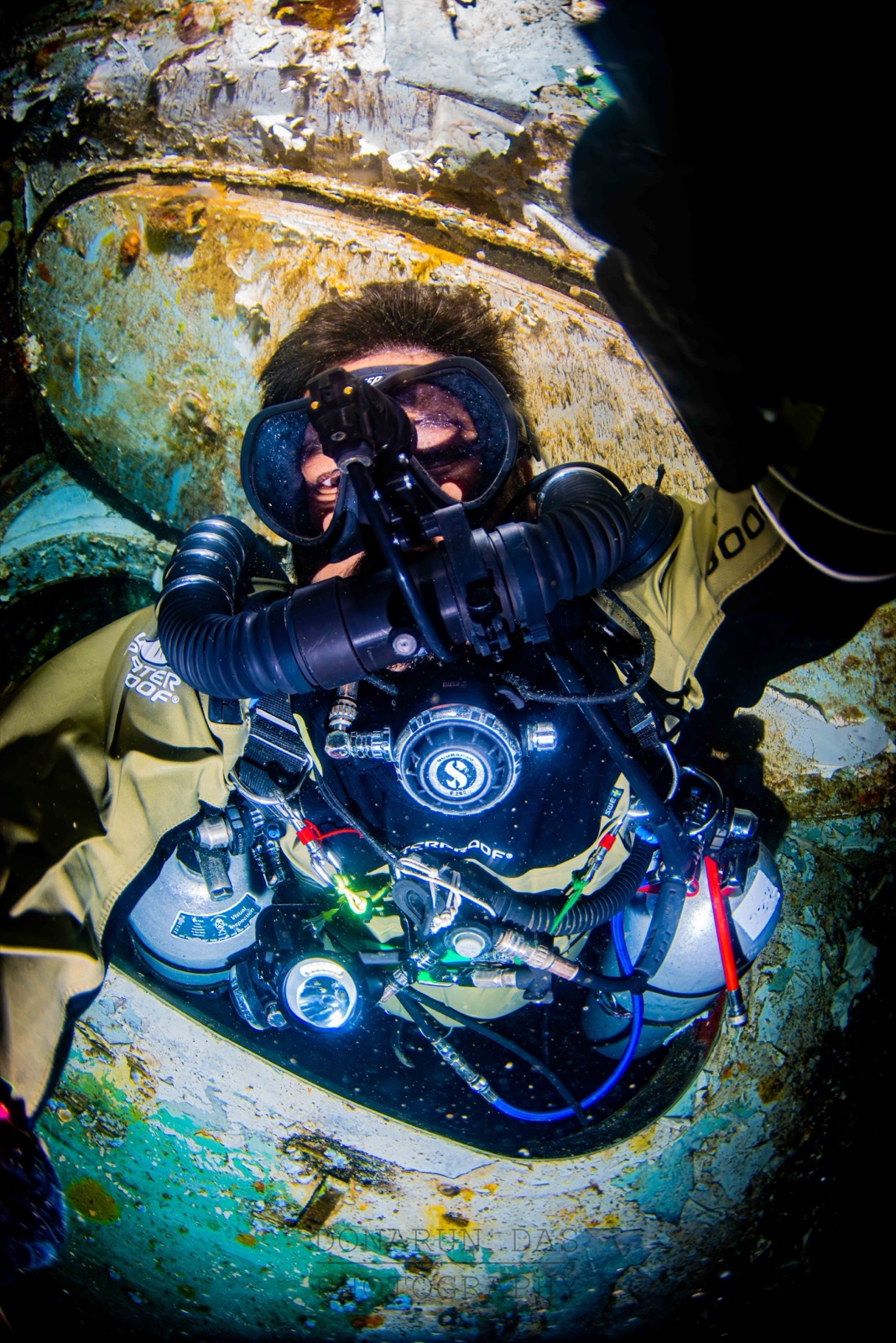 CCR diver inside the rusted opening of a steel wreck, breathing loop over the shoulders, facing the camera
