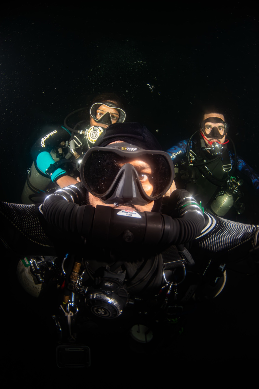 Three divers grouped close in dark water — a CCR diver on a chest-mounted loop in front, two backmount divers lit behind